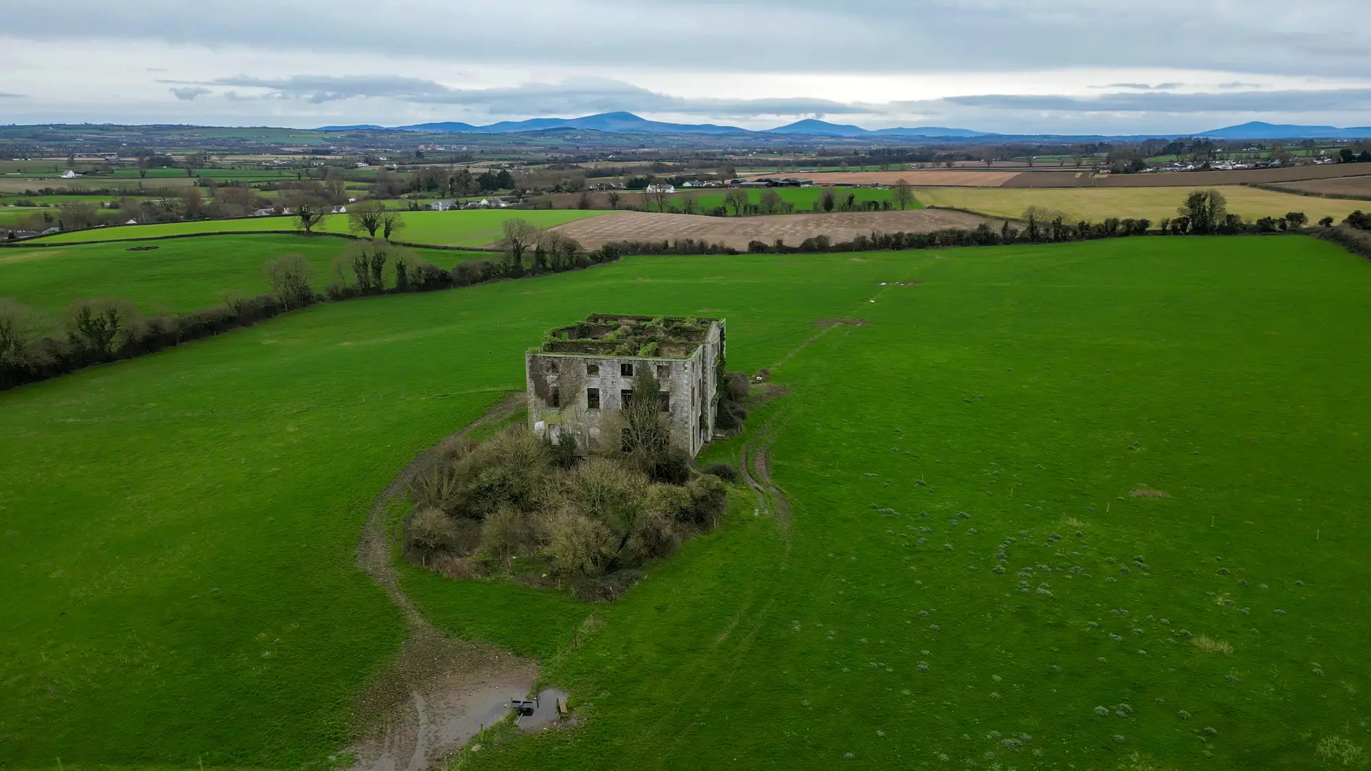 A roofless stone tower house standing alone in a green field, seen from a drone